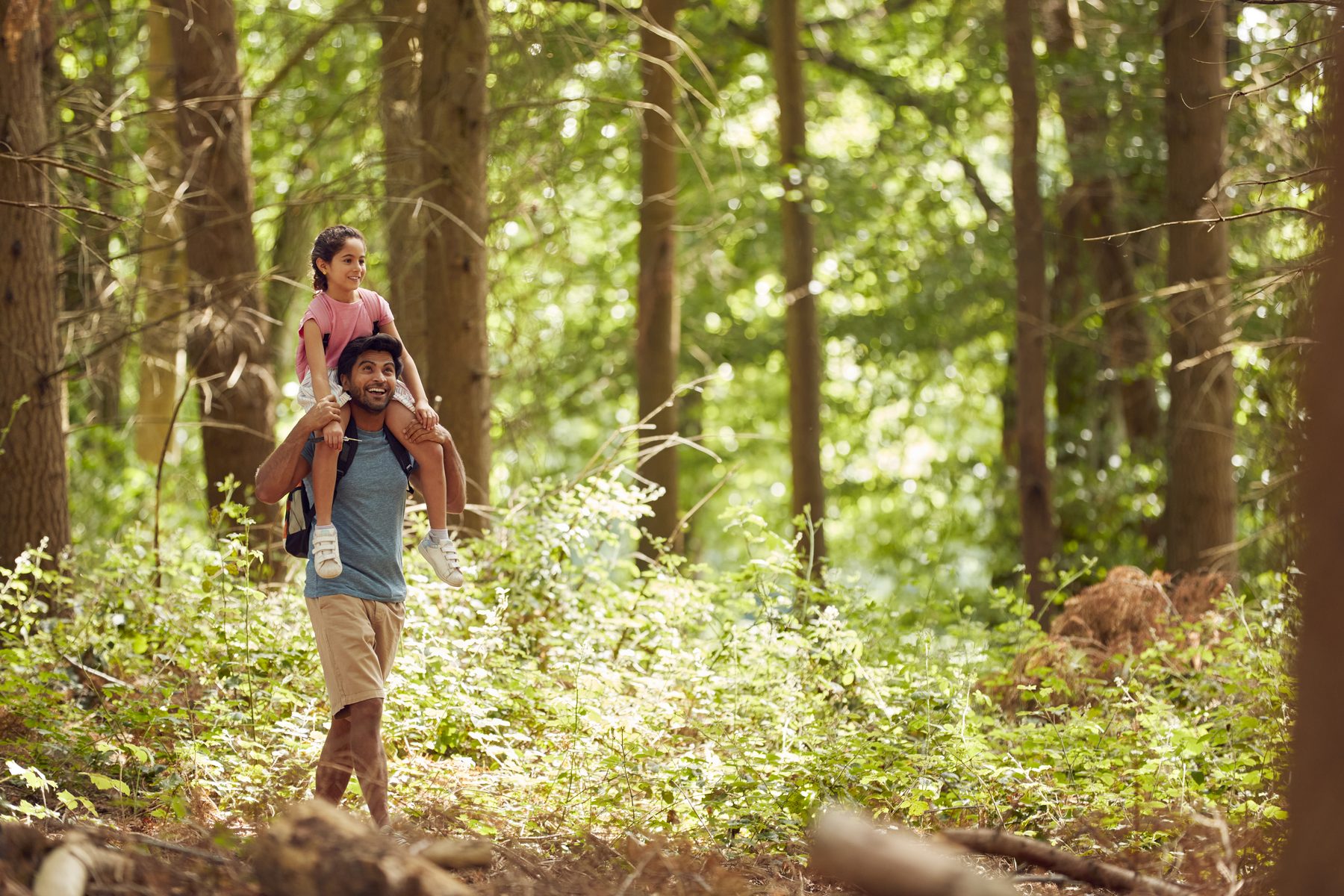 A father carrying his daughter on his shoulders through a sunlit British wood